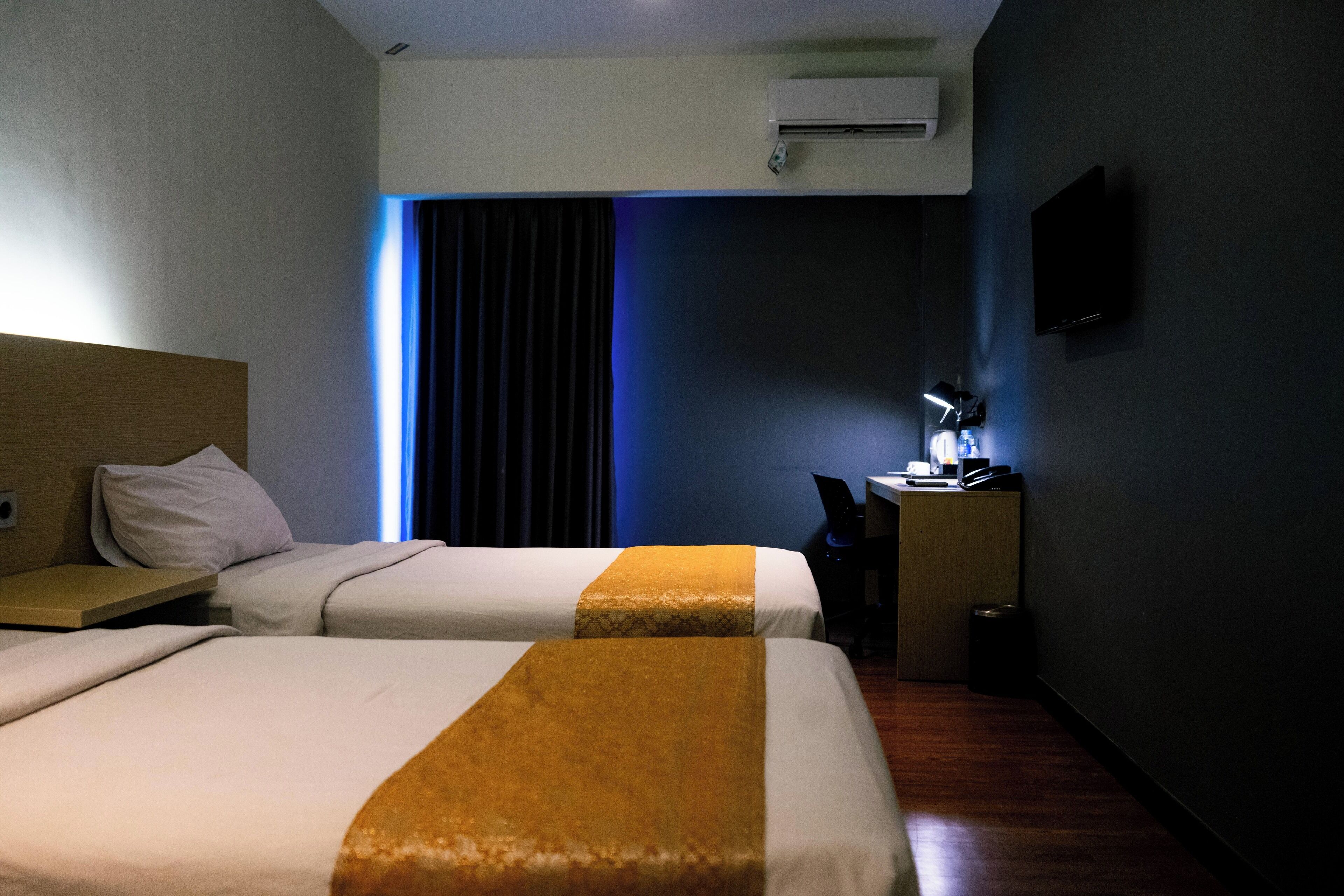 A hotel room with two beds, a desk with a chair, a TV mounted on the wall, and an air conditioning unit.