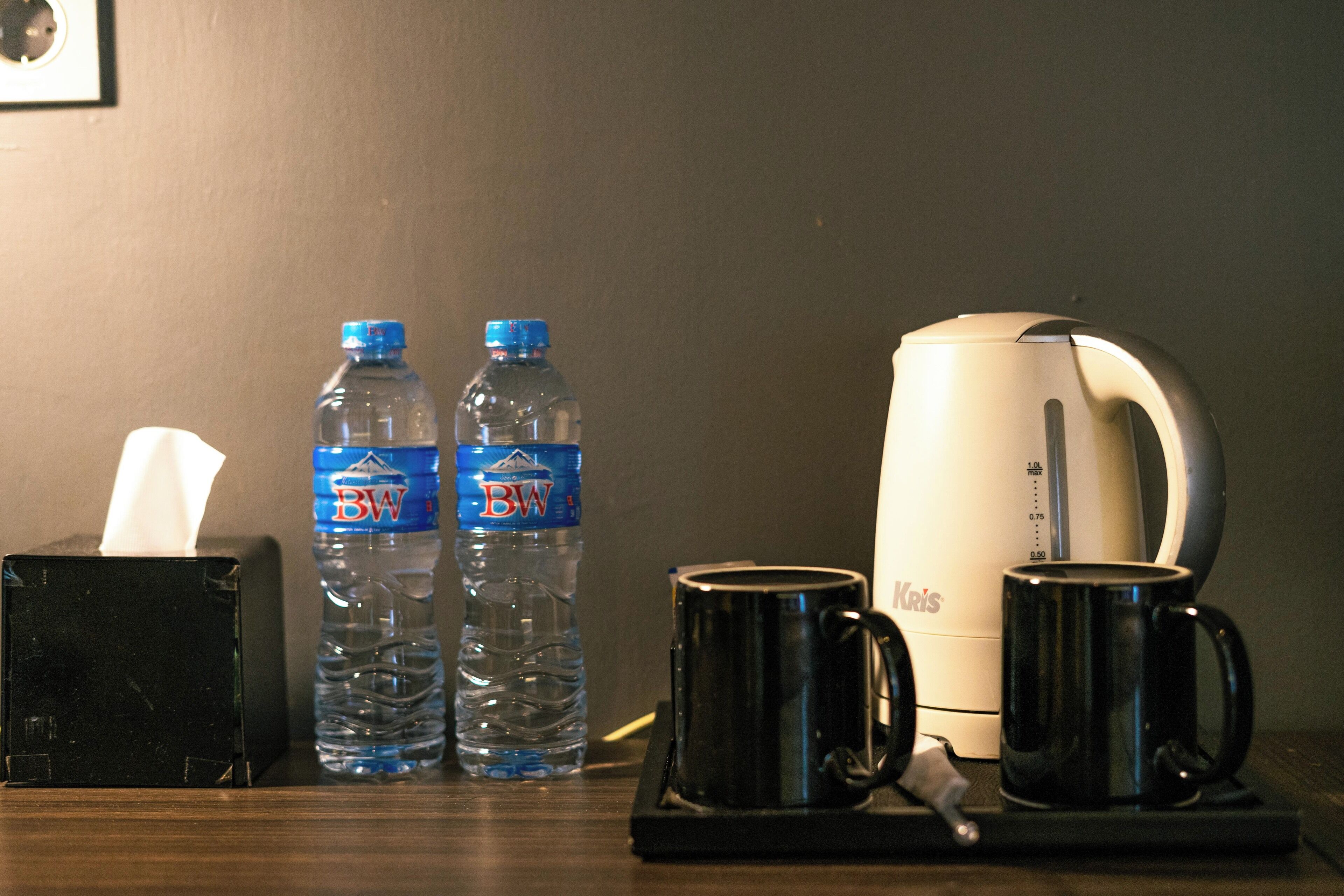 A table with a tissue box, two water bottles, a coffee maker, and two mugs.