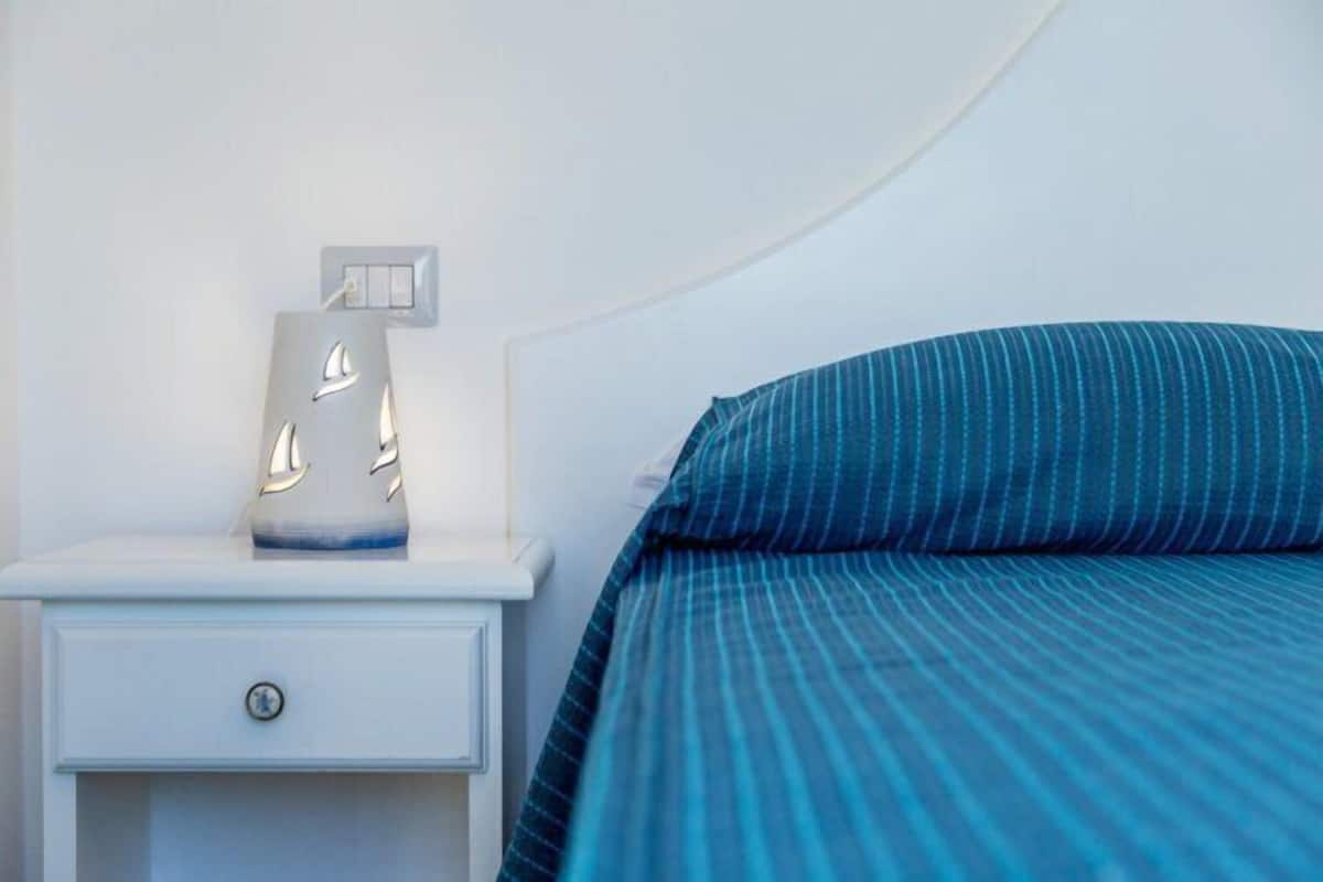 Una cama bien tendida con una colcha azul a rayas y una mesita de noche blanca con una lámpara.