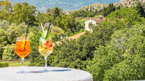 A table with two glasses of drink, a view of a mountainous landscape, and a building with a tiled roof. - Hotel Agugliastra (Baunei)