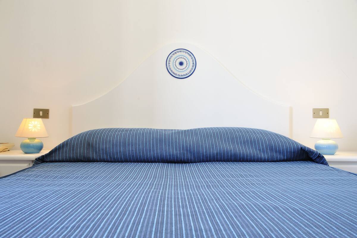 Una cama bien hecha con una colcha azul a rayas y un cojín a juego, flanqueada por dos lámparas de noche sobre mesitas blancas.