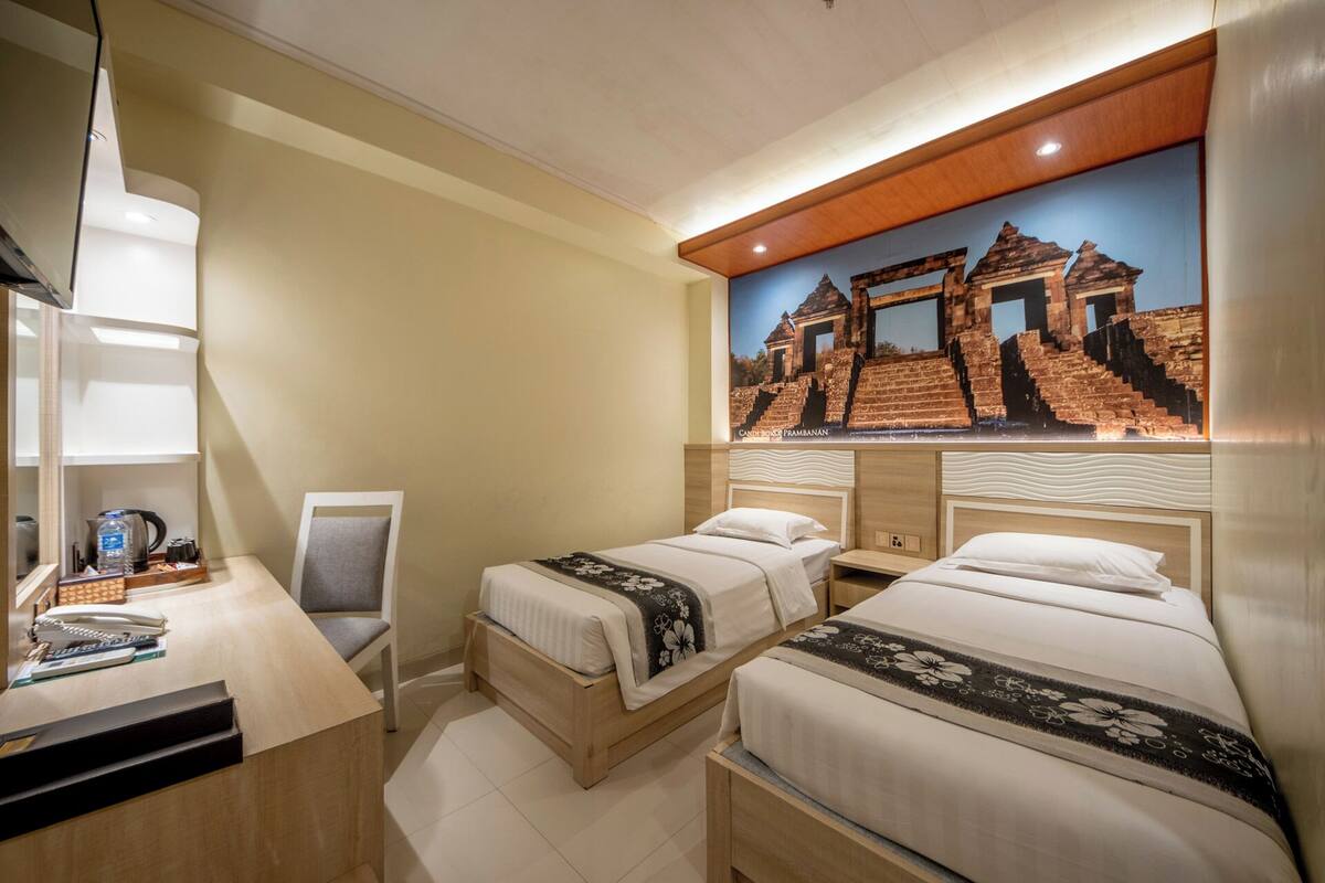 A hotel room with two beds, a desk with a telephone and a water bottle, a chair, and a wall-mounted picture of an ancient structure.