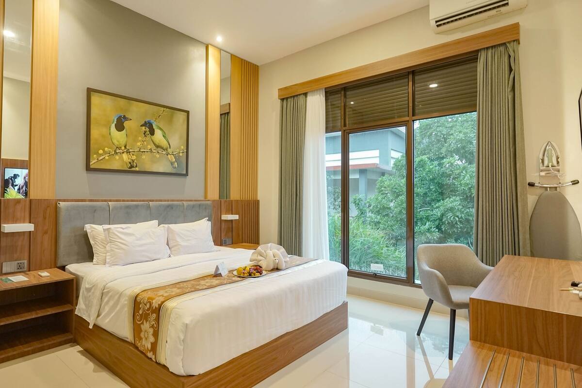 A modern hotel room with a large bed, a desk, and a view of greenery outside.