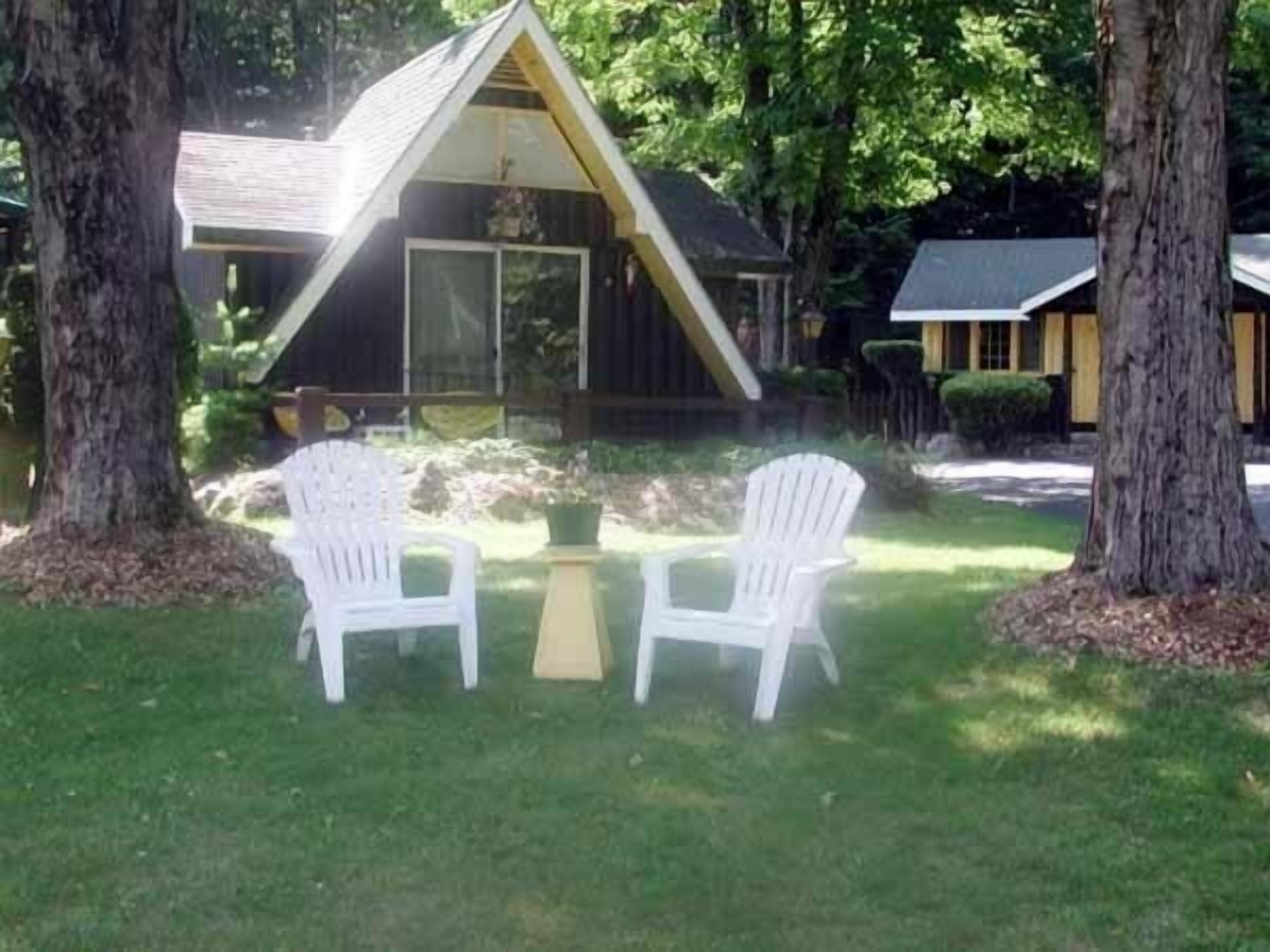 A small, triangular-shaped building with a porch, two white plastic chairs, and a table in front.