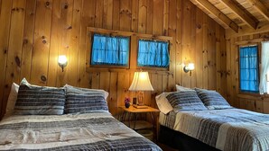 A wooden cabin room with two beds, a small table, and two windows with curtains. - Amber Lantern Lake George Motel and Cottage Rentals (Lake George)
