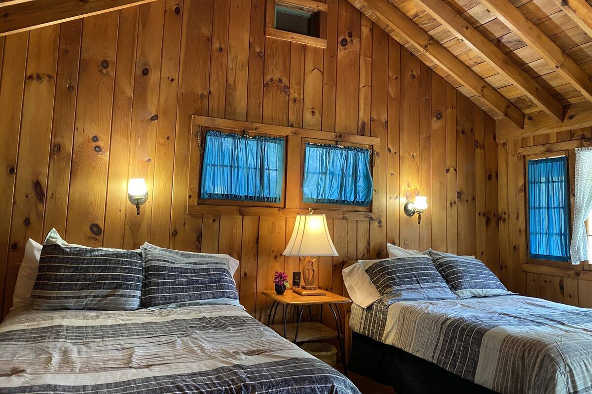 A wooden cabin room with two beds, a small table, and two windows with curtains.