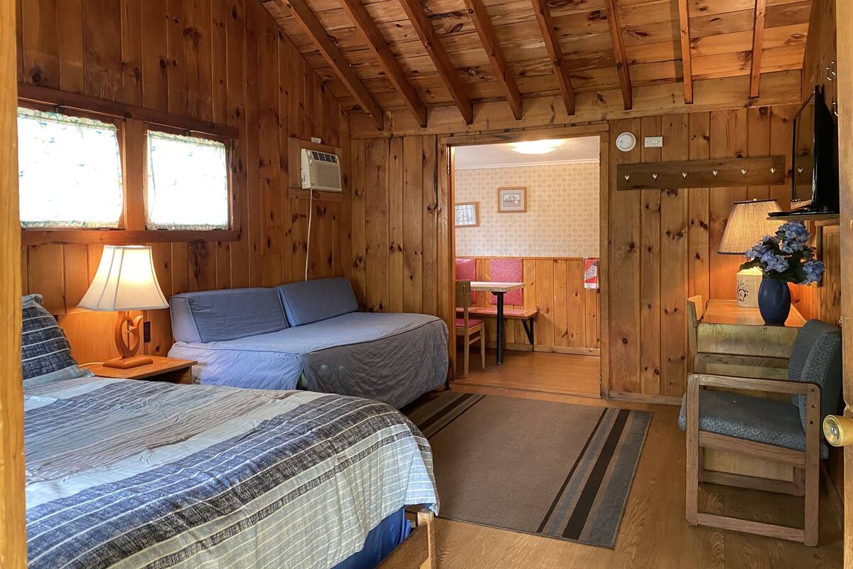 A wooden cabin room with two beds, a table, a chair, and a small kitchenette area.