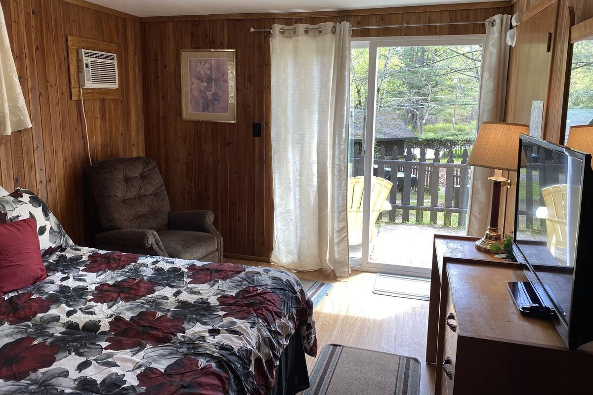 A bedroom with a bed, a chair, a desk with a lamp, a window with curtains, and a view of the outside.
