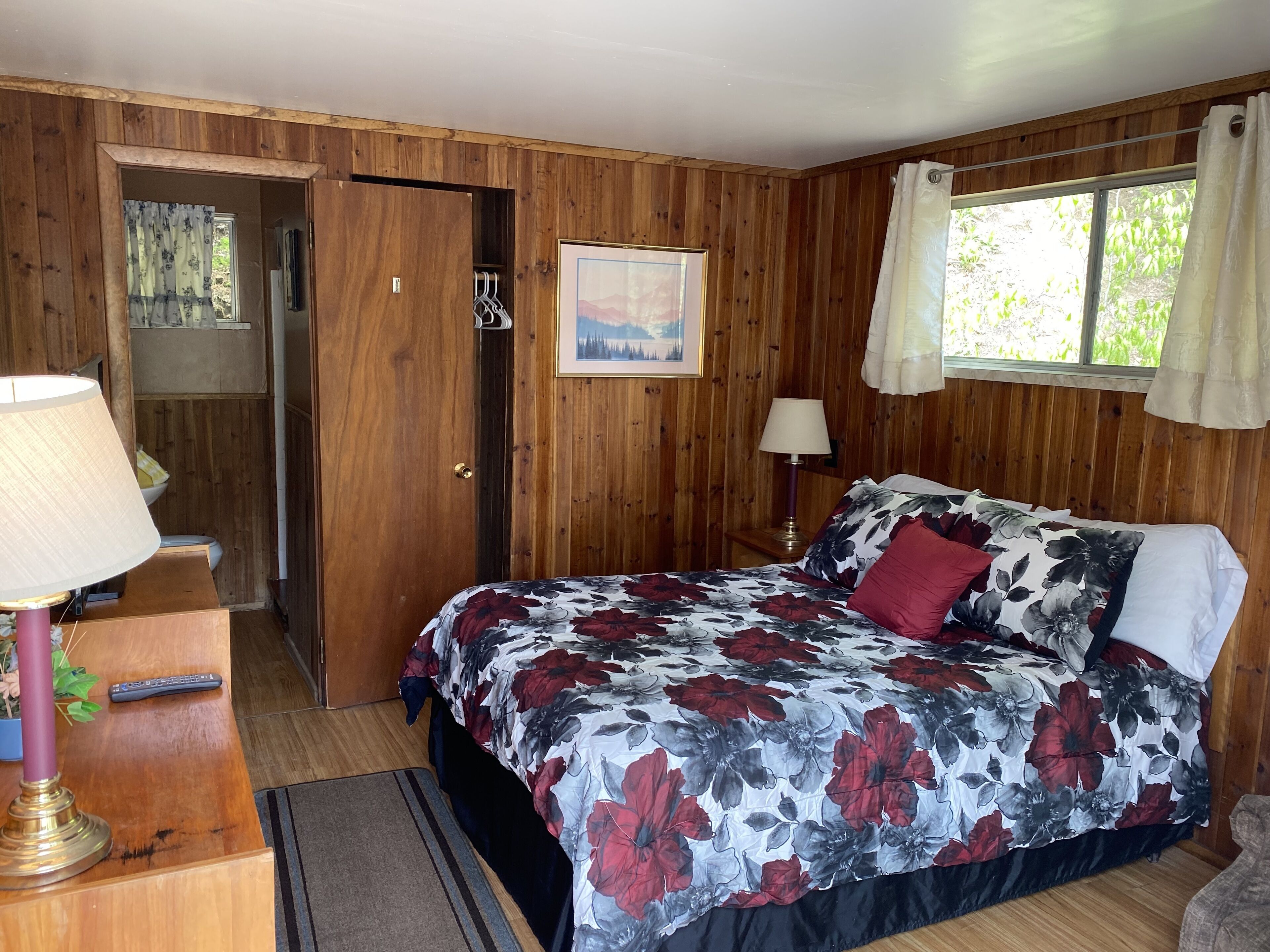 A wooden-paneled bedroom with a bed, two bedside lamps, a painting, and a window with curtains.