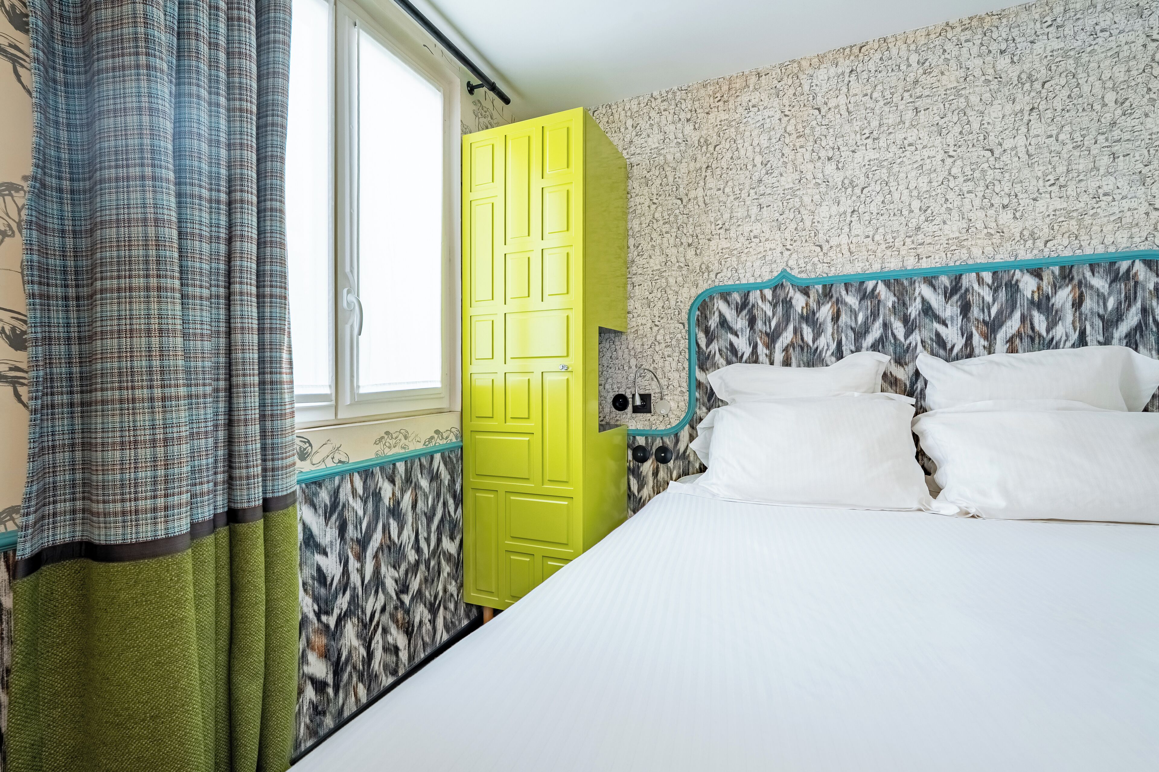 A room with a bed, a yellow door, patterned wallpaper, and a window.