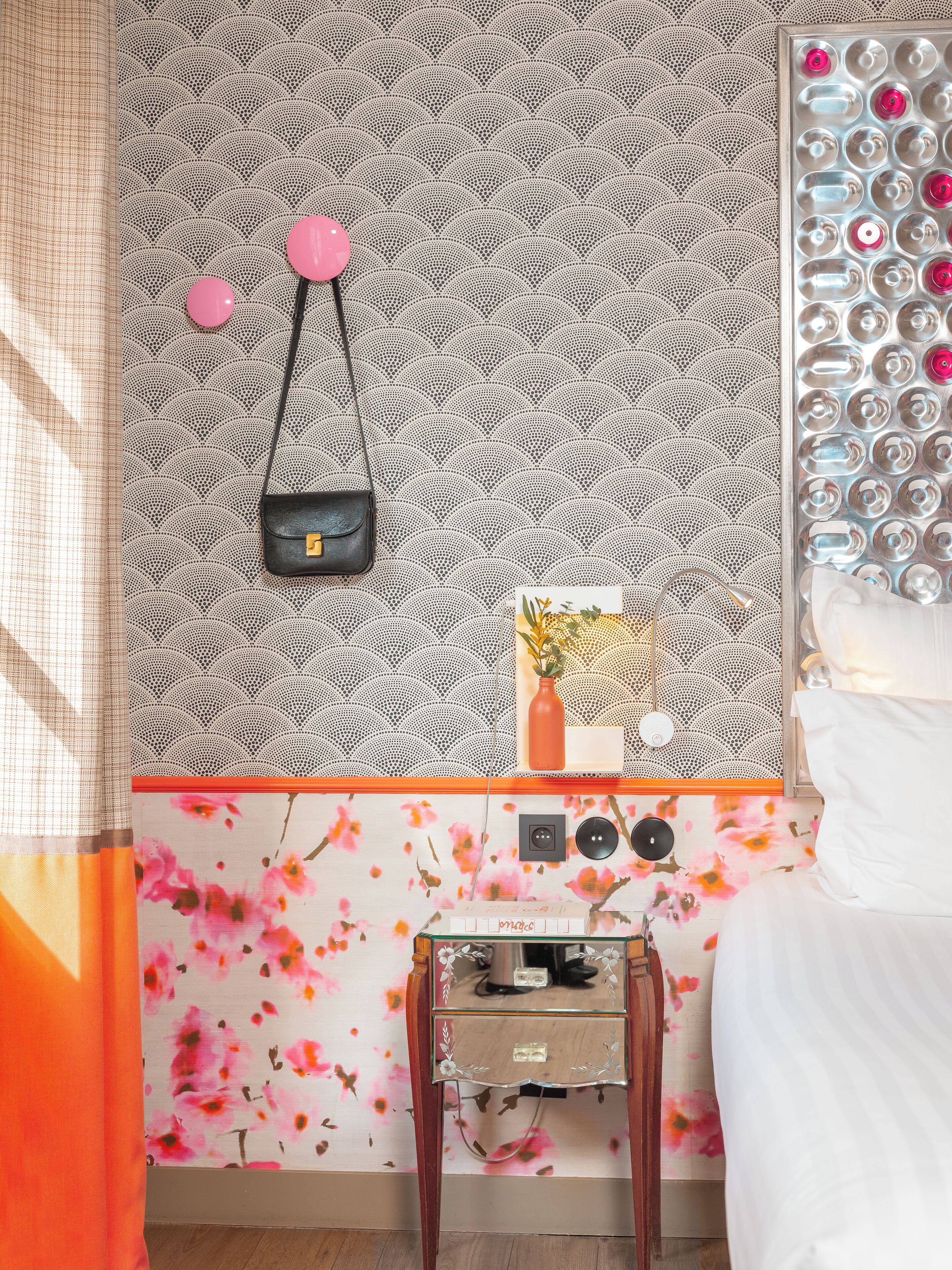 A bedroom with a patterned wallpaper, a small bedside table with a vase, a power outlet, and a lamp.