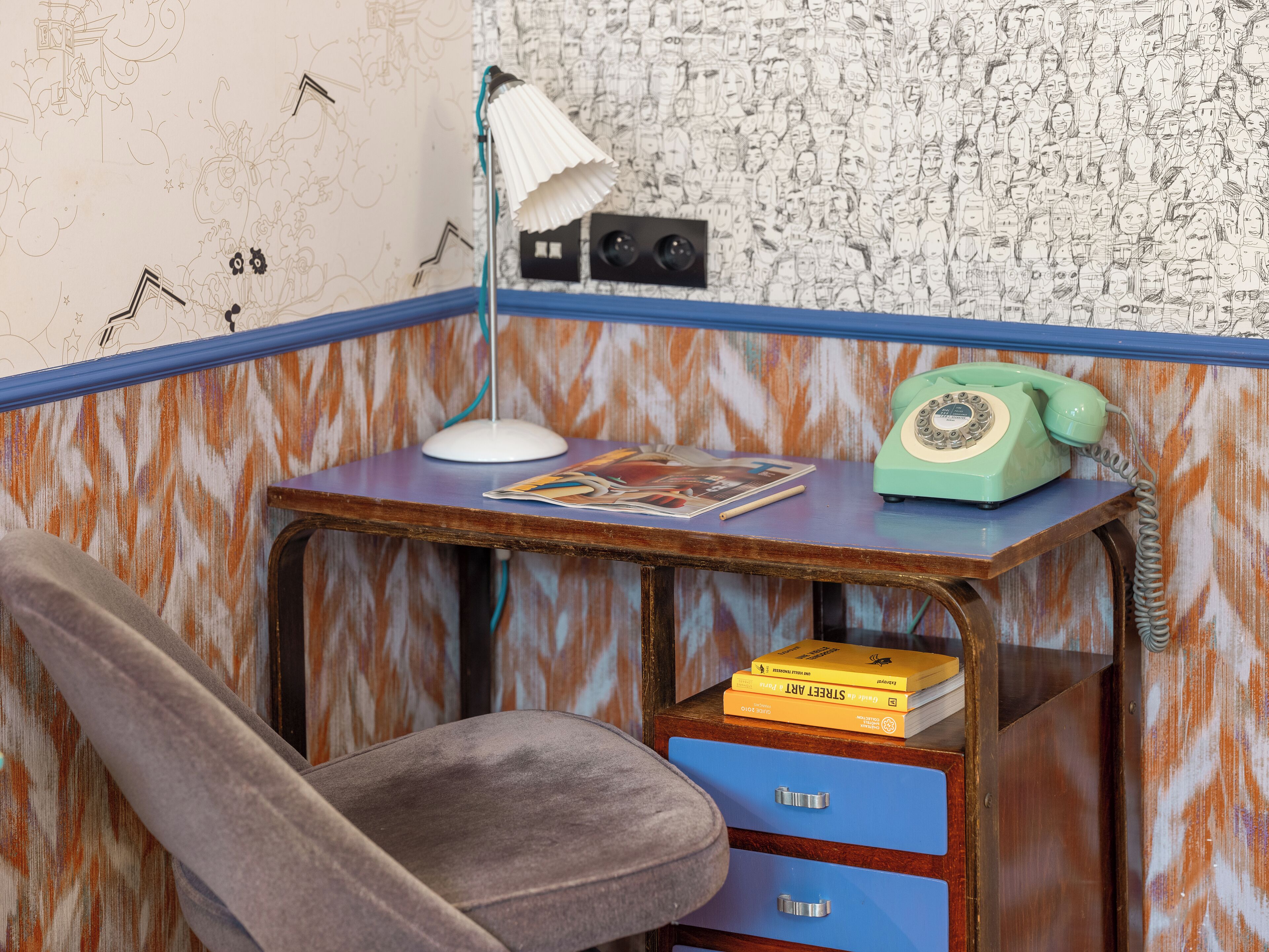Een hoek van een kamer met een bureau, een stoel, een lamp, een telefoon en een wand met een patroon.