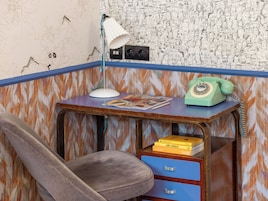 A corner of a room with a desk, a chair, a lamp, a telephone, and a wall with a patterned design.