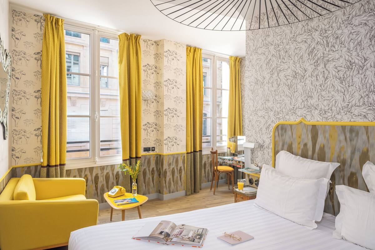 A hotel room with a bed, a yellow armchair, a small table with a lamp, and a window with yellow curtains.