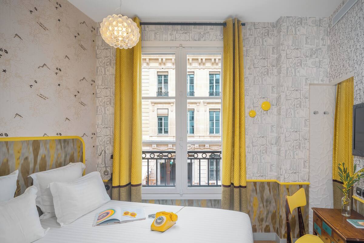 A hotel room with a bed, a desk, a chair, a telephone, and a window with yellow curtains.