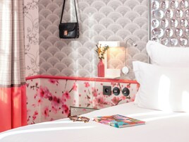Ein ordentlich bezogenes Bett mit einer Zeitschrift und Sonnenbrille, ein kleiner Tisch mit einer Vase und einer Lampe sowie eine Wand mit dekorativem Muster.