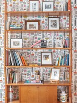A bookshelf with various books and framed pictures against a patterned wallpaper background.