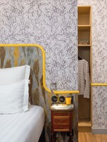 Un dormitorio con cama, una mesita de noche con lámpara, una estantería con libros y un armario con ropa colgada.