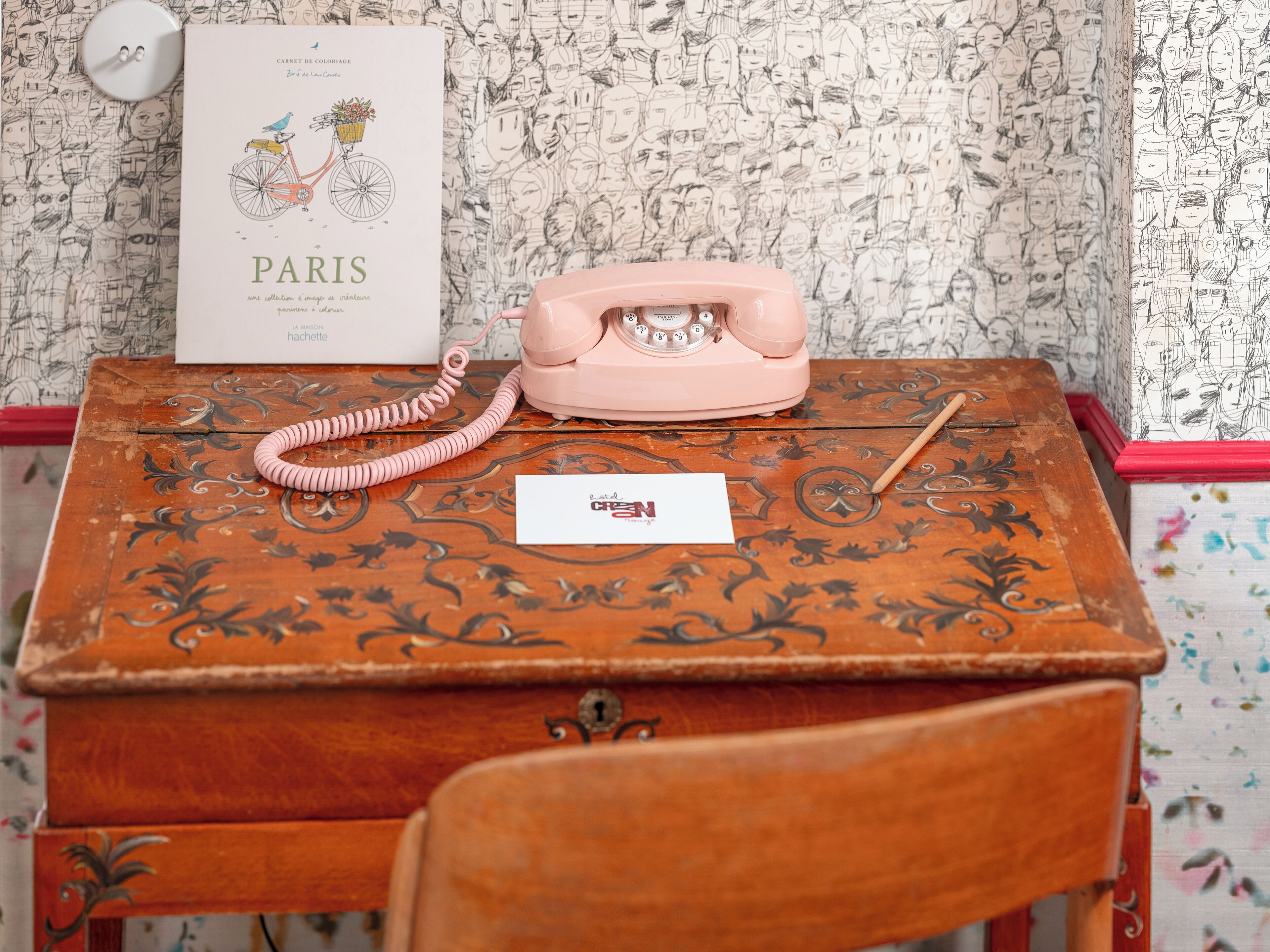 Un teléfono antiguo sobre un escritorio de madera con papel tapiz estampado, una postal y una pluma.