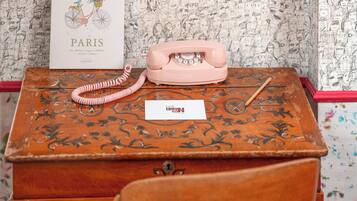 Un teléfono antiguo sobre un escritorio de madera con papel tapiz estampado, una postal y una pluma.