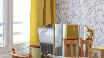 A room with a glass table, two wooden chairs, a yellow cushion, a yellow curtain, and a window with a view.