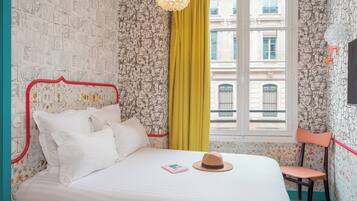 A neatly made bed with white linens and a pink book, a yellow curtain, and a window with a view of buildings.