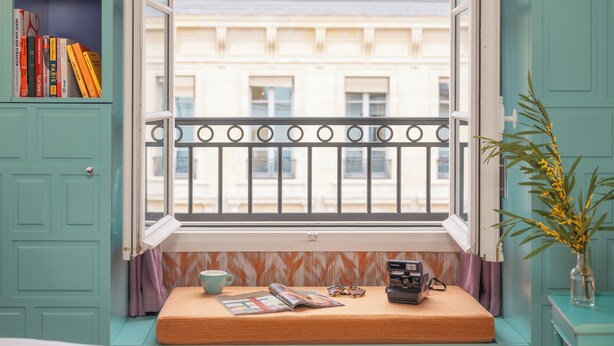 A room with a window overlooking a building, a bookshelf with books, a small bench with items, and a plant. - Hôtel Crayon Rouge (Paris)