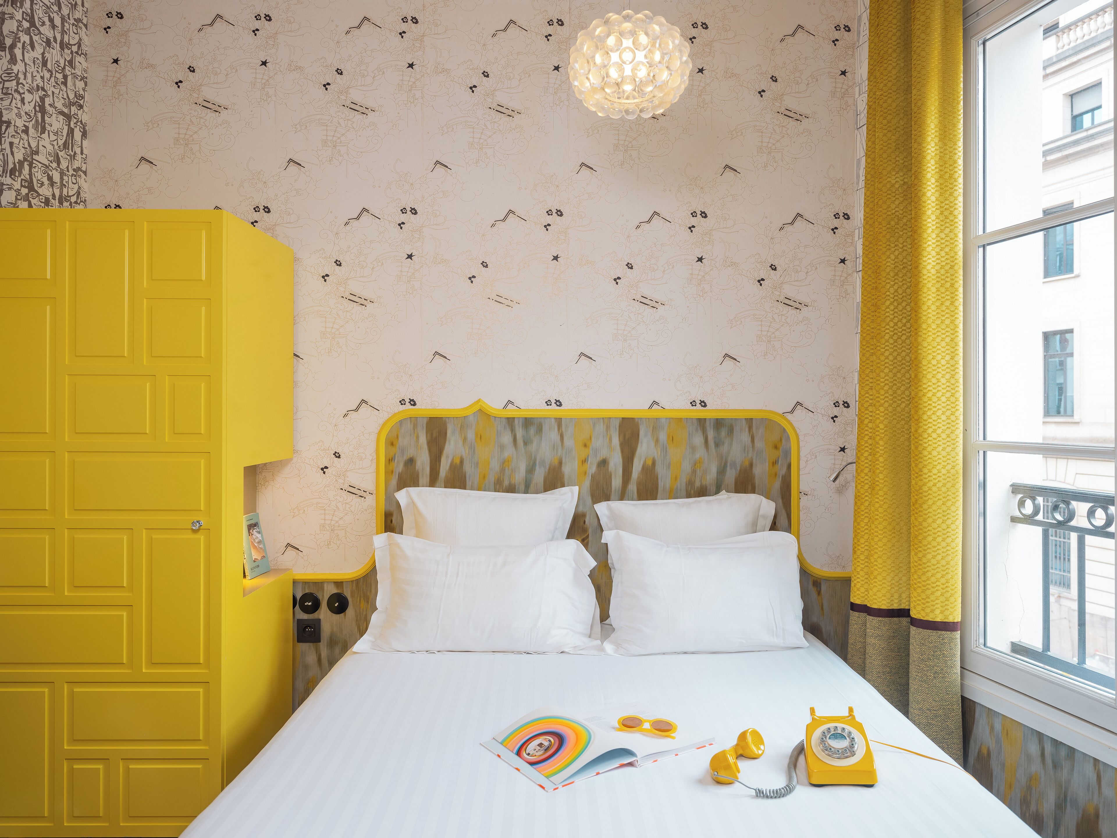 A room with a bed, a yellow wardrobe, a patterned wallpaper, and a window with curtains.