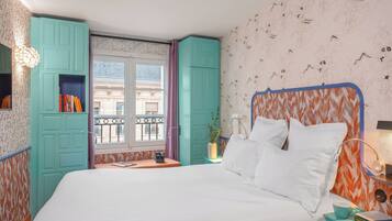 Una cama bien hecha con ropa blanca, una mesita de noche color turquesa con una taza y libros, y una ventana con vista a edificios.