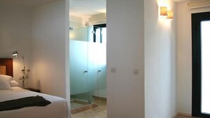 Una habitación de hotel con cama, escritorio, lámpara, cabina de ducha de vidrio y puerta que da a otra habitación.