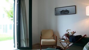 Una habitación con una silla, una mesa con un florero y un cuadro en la pared, y vistas al exterior con vegetación.