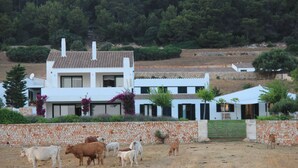 Un edificio blanco con pared de piedra, rodeado por un campo seco y árboles.