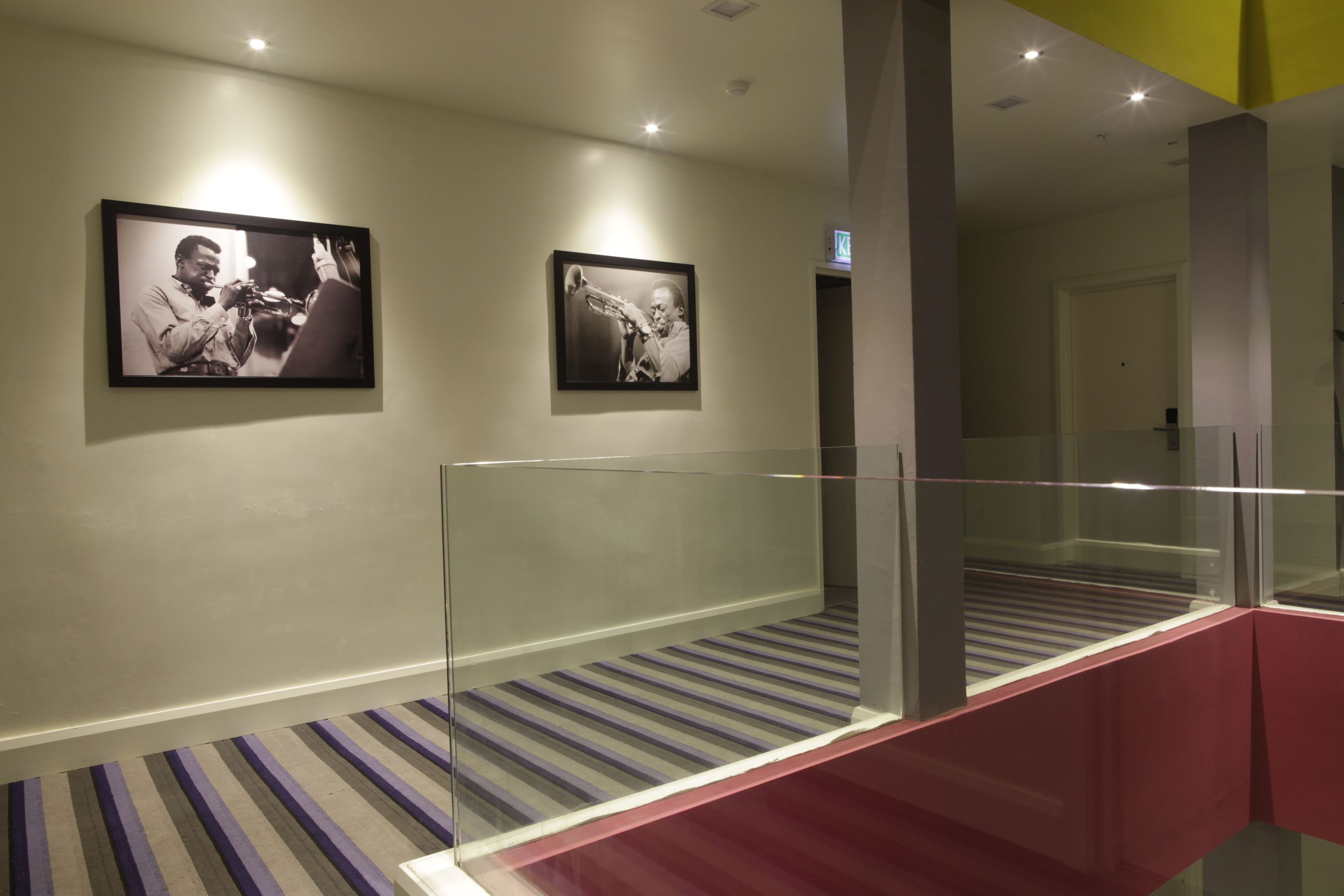 Un pasillo con dos fotografías en blanco y negro enmarcadas en la pared, un suelo de alfombra a rayas y una barandilla de vidrio en una escalera roja.
