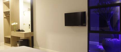 A modern hotel room with a large bed, a vanity with a mirror, a flat-screen TV mounted on the wall, and a large window with a view of a decorative planter.