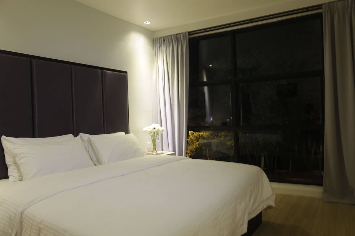 Una cama bien hecha con sábanas blancas y un cabecero oscuro en una habitación con un amplio ventanal y cortinas.