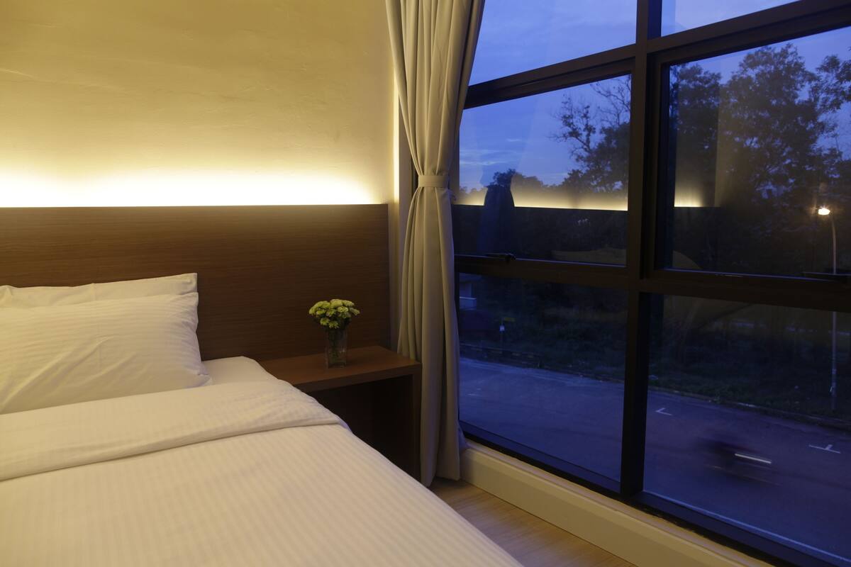 Habitación de hotel con un ventanal grande, una cama con sábanas blancas, una mesita de noche con un jarrón de flores y vistas al cielo nocturno.