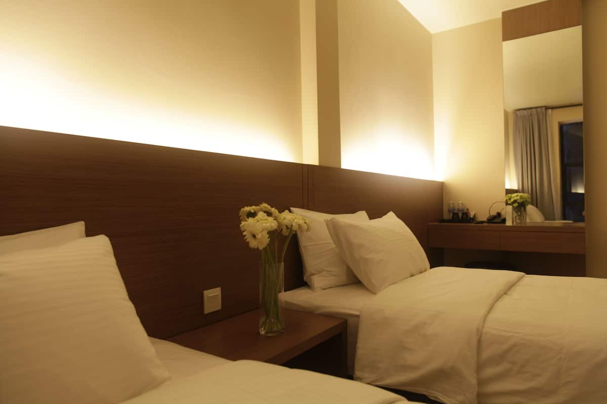 Habitación de hotel con dos camas, una mesita de noche, un florero con flores, un espejo y un cabecero de madera.