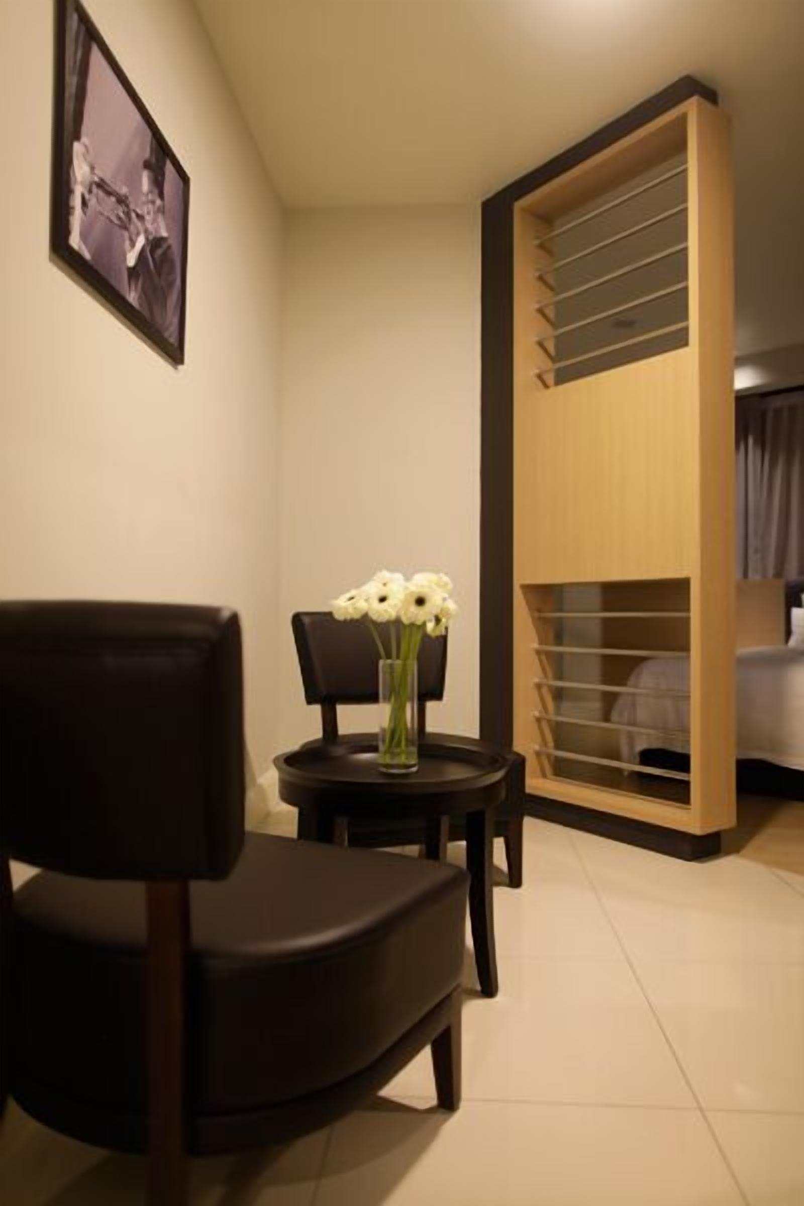 A hotel room with a bed, a wooden wardrobe with multiple shelves, a small table with a vase of flowers, a chair, and a framed picture on the wall.