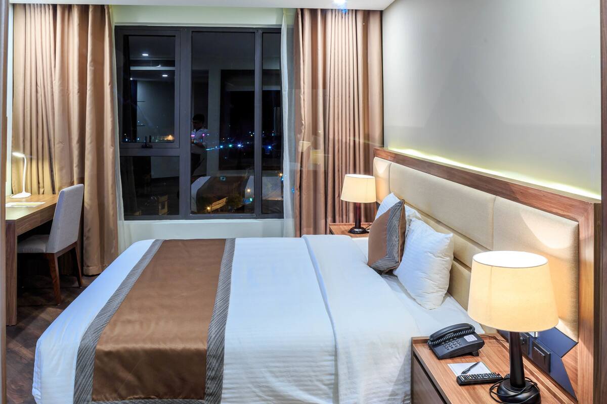 A hotel room with a bed, bedside tables, a desk, and a view of the city at night.