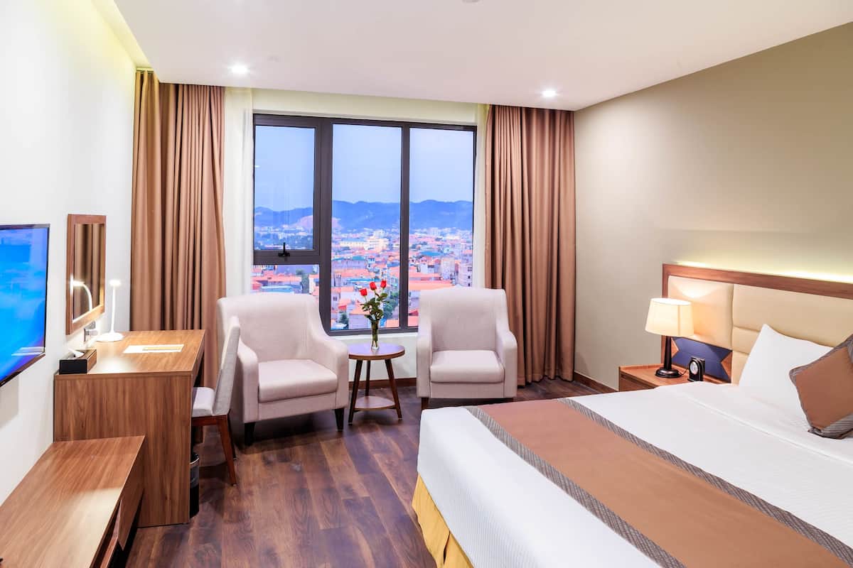 A hotel room with a large window, a bed, two armchairs, a desk, and a TV.