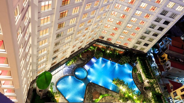 A multi-story hotel with a rooftop pool area featuring a circular water feature and surrounding greenery.