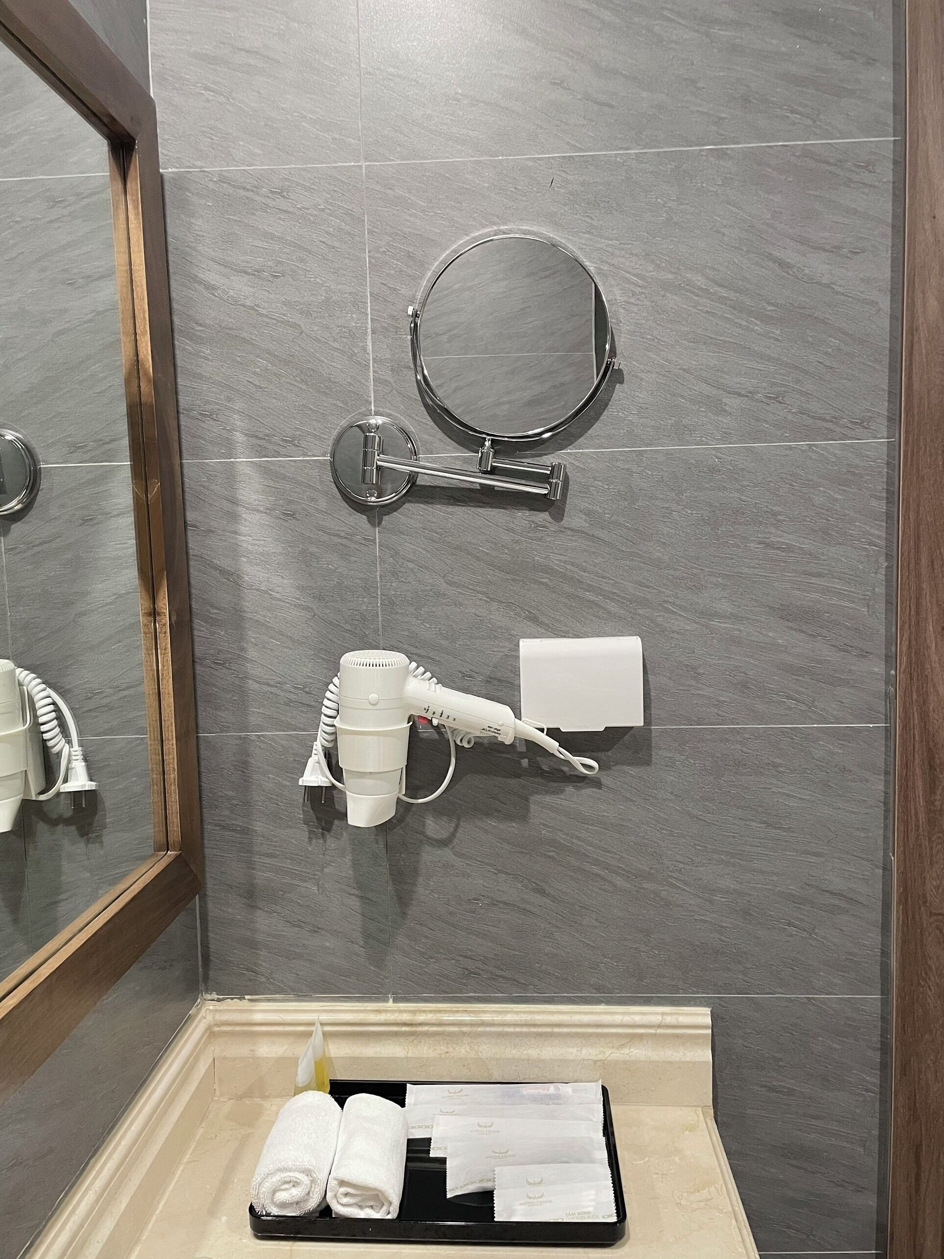 A bathroom with grey tiles, a mirror, a towel rack, a hairdryer, and a tray with towels and toiletries.