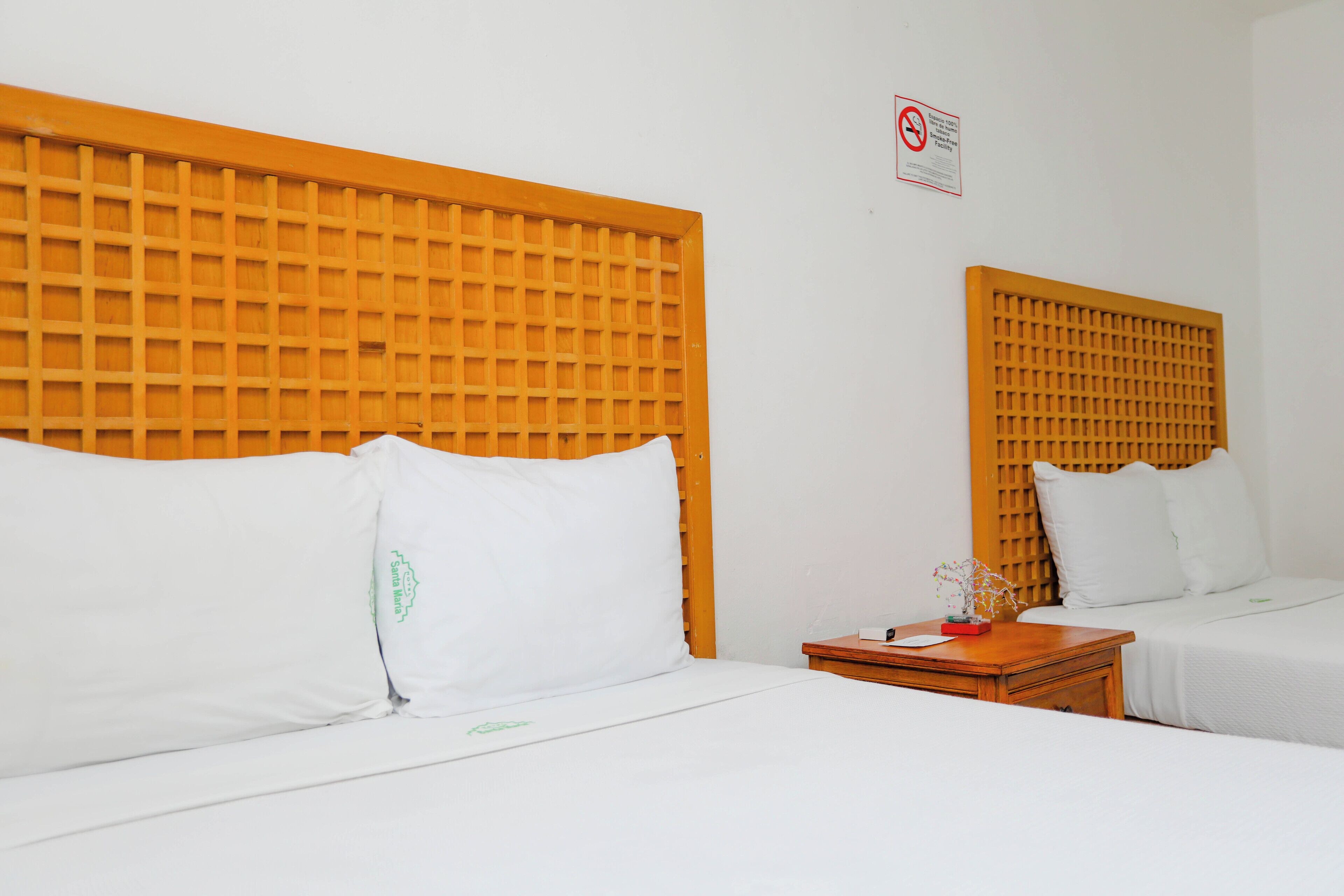 A hotel room with two beds, each with a wooden headboard and white bedding. There is a small wooden nightstand with a lamp and a flower vase between the beds.
