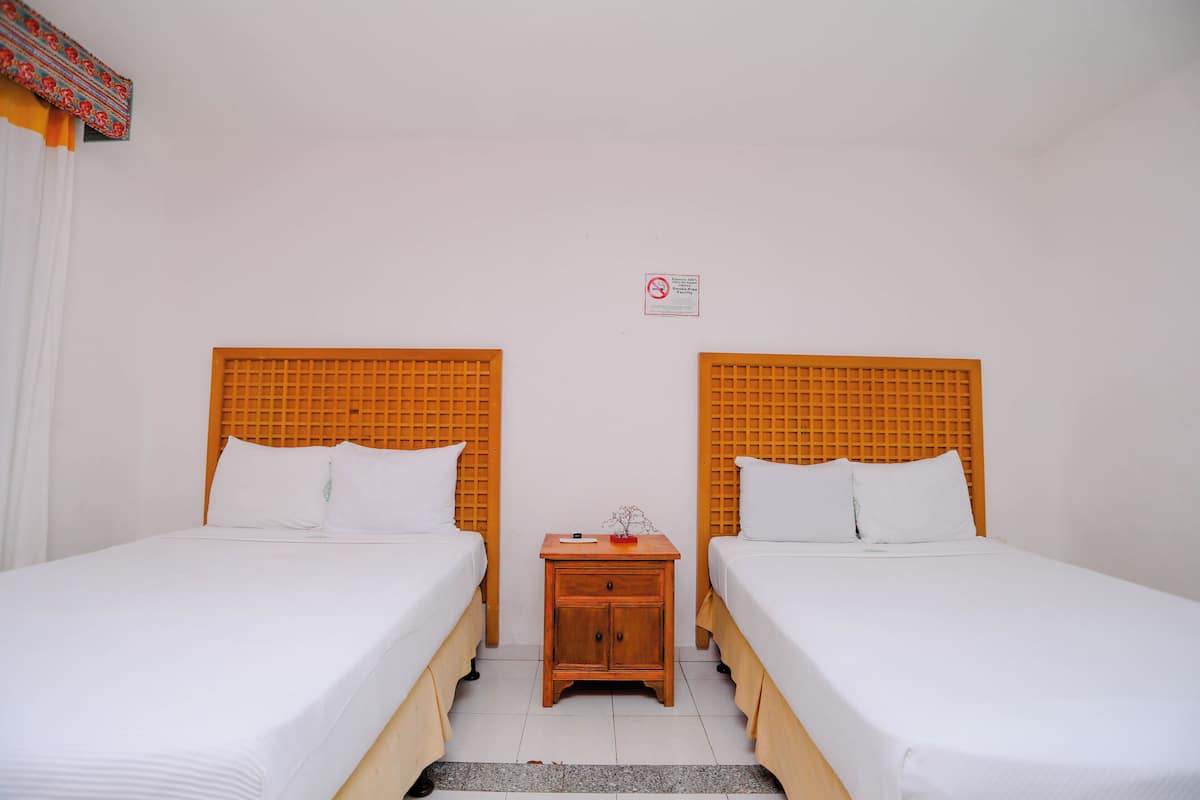Una camera d'albergo con due letti singoli, un comodino in legno con un piccolo tavolo e una parete bianca con il cartello ‘vietato fumare.’