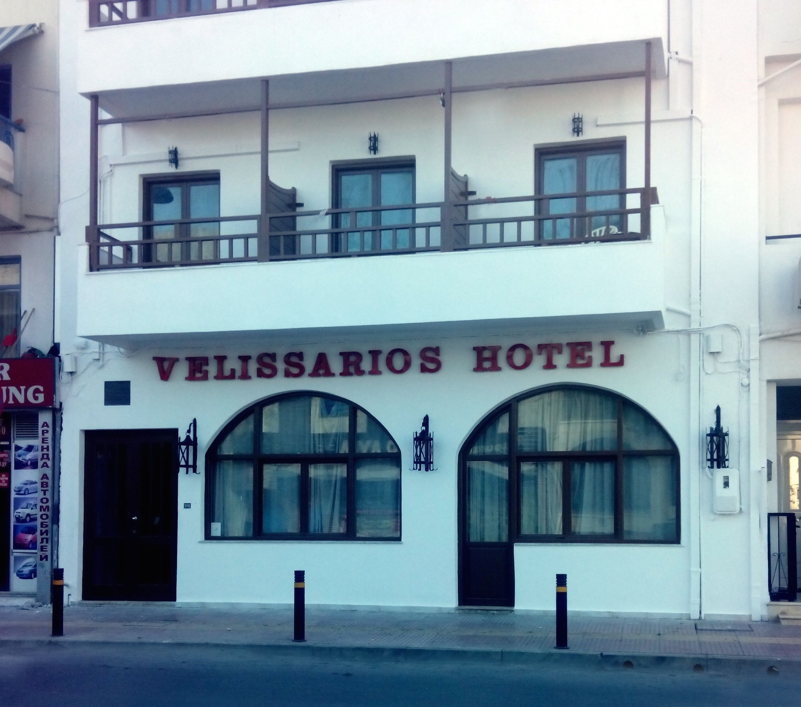 A two-story building with the sign "VELISSARIOS HOTEL" above the entrance. There are two large arched windows on the ground floor and a balcony on the upper floor.