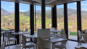 Een eethoek met tafels en stoelen, grote ramen met uitzicht op bergen en velden, en een modern plafondontwerp.
