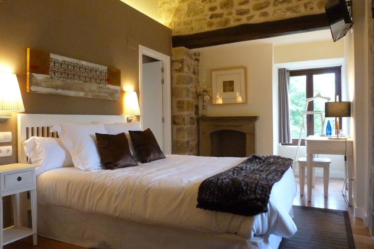 A bedroom with a stone wall, a fireplace, a bed with white bedding, a nightstand, and a desk with a lamp.