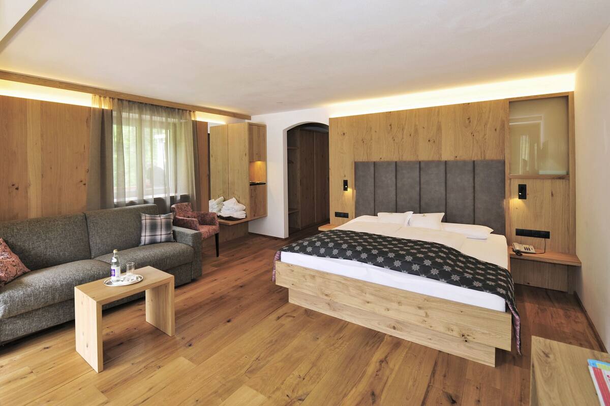A modern hotel room with a large bed, a sofa, a small table, and wooden paneling.