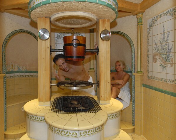 A sauna with a central heating element, a tiled wall with a floral pattern, and a wooden ceiling.
