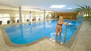 Una piscina interior con agua cristalina azul, suelo de baldosas y una persona de pie junto a la piscina.